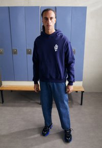 Marineblå hettegenser med frontlomme og hvit grafikk, paret med avslappede blå jeans og sorte sneakers med blå detaljer, satt i et garderoberom.