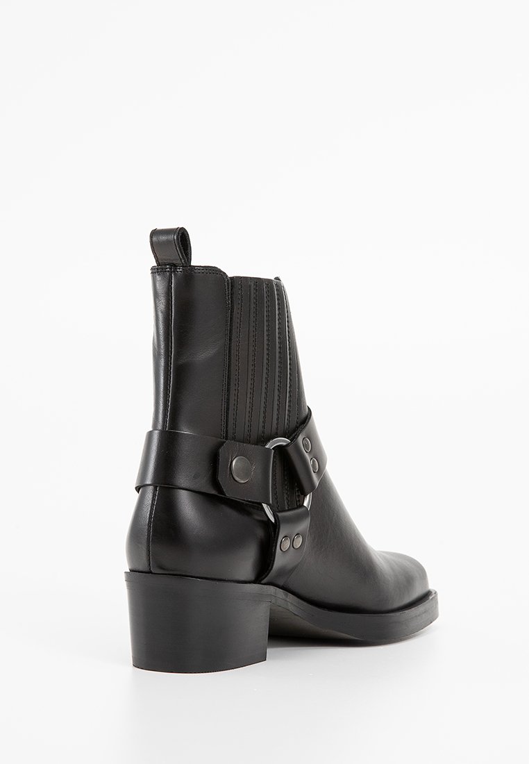 Stivaletto in pelle nera con una forma curva, pannelli a coste, una fascia ampia e un dettaglio con fibbia, e un tacco a blocco basso. Texture liscia complessivamente.