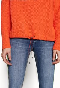 Suéter de color naranja con textura, de manga larga y con dobladillo ajustable, combinado con jeans azules que tienen llamativos adornos laterales en amarillo brillante.