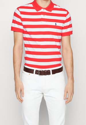 Mand iført rød og hvid vandret striped poloshirt med lille blå logo, hvide bukser og et mørkebrunt bælte.