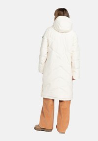 Manteau doudoune long blanc avec capuche, présentant un motif matelassé en chevrons. Associé à un pantalon en velours côtelé orange et des bottes marron.