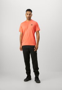 Tricou sportiv portocaliu cu mâneci scurte și un logo negru. Asortat cu pantaloni scurți negri, completând un look sportiv.