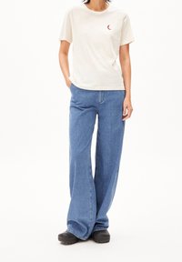 Beige bomull t-shirt med en liten röd halvmåne-design, paret med blå vida jeans och svarta slip-on skor. Modellen har händerna i fickorna.