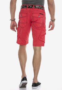 Rote Cargo-Shorts mit mehreren Taschen, sichtbaren Nähten und einem schwarzen Gürtel mit silbernen Nieten. Tragen mit schwarzen Sneakers.