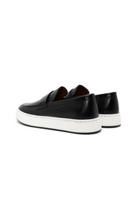 Schwarze Lederloafers mit weißen, dicken Gummisohlen, von hinten aus einem leichten Winkel auf einem weißen Hintergrund betrachtet.