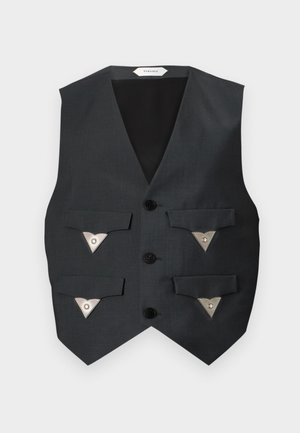 Fiatal, göndör hajú férfi, aki ujjatlan fekete mellényt visel háromszög alakú gombokkal, bő, csíkos nadrágot, barna cipőket és egy medálos nyakláncot.