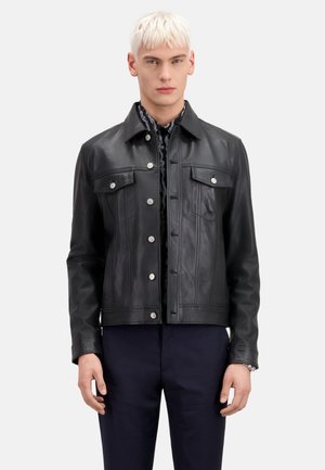 Chaqueta de cuero negra con un diseño cropped, que presenta dos bolsillos en el pecho, botones a presión y detalles de costuras verticales. Lleva puesta una camisa con estampado.