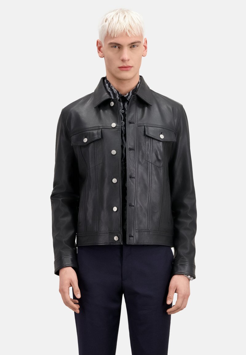 Giacca in pelle nera con design corto, caratterizzata da due tasche sul petto, bottoni a scatto e dettagli di cucitura verticale. Indossata sopra una camicia con fantasia.