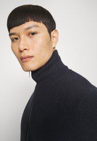 Marineblauer, gerippter Rollkragenpullover mit Zip-Verschluss, strukturiertem Stoff und taillierter Passform. Nahaufnahme, die den Kragen und die Stitch-Details hervorhebt.