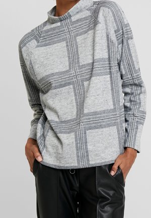 Personne portant un pull gris à manches longues avec de grands motifs à carreaux noirs et un pantalon en cuir noir, mains dans les poches.