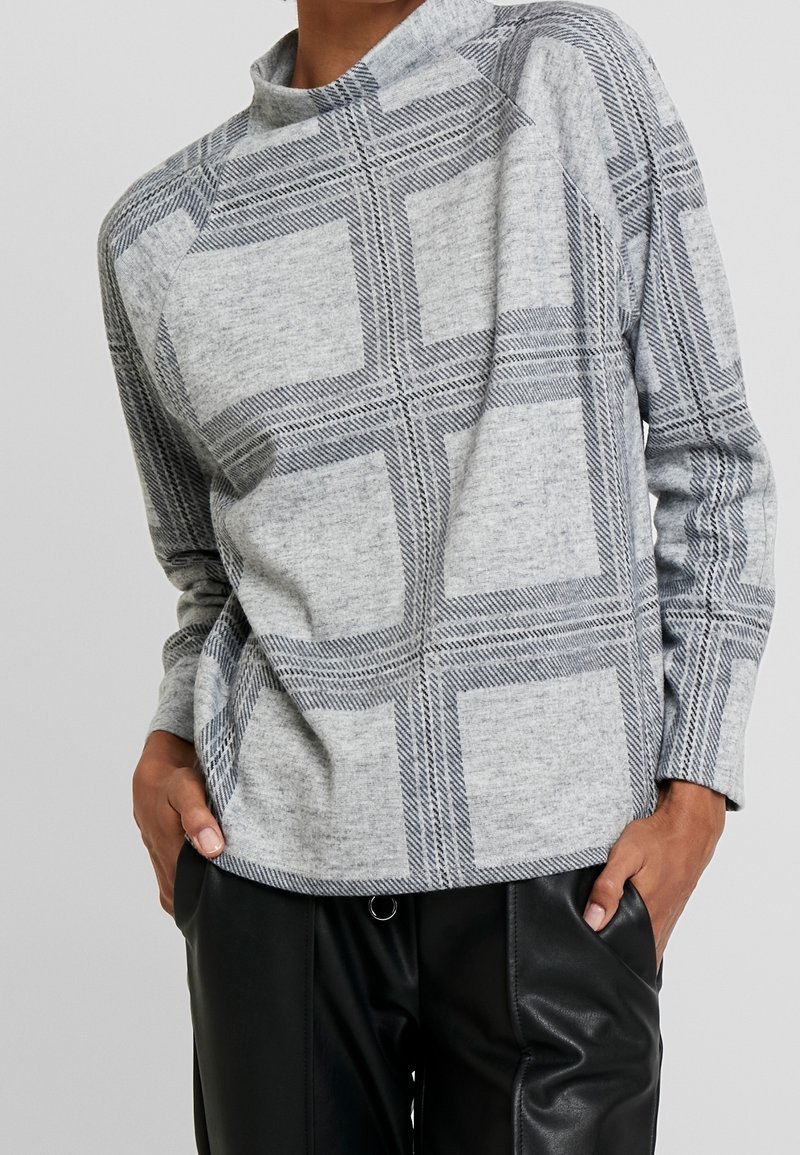 Personne portant un pull gris à manches longues avec de grands motifs à carreaux noirs et un pantalon en cuir noir, mains dans les poches.