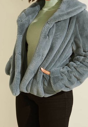 Femme portant une veste en fausse fourrure bleu clair sur un pull à col roulé vert avec un pantalon noir, main glissée dans la poche de la veste.