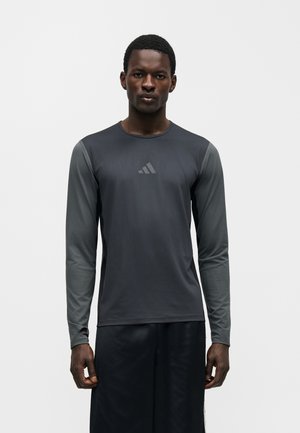 Homme debout, face à l’avant, portant un tee-shirt de sport ajusté gris foncé à manches longues avec un logo discret sur la poitrine, sur un fond clair uni.