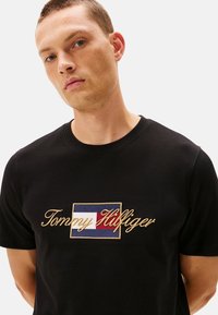 Camiseta de algodón negra con el logotipo "Tommy Hilfiger" bordado en dorado y un parche rectangular en colores azul, blanco y rojo.