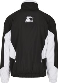 Chaqueta cortavientos en blanco y negro con un alto cuello, puños elásticos y cinturilla. Presenta un logo en la parte trasera y paneles laterales en contraste.