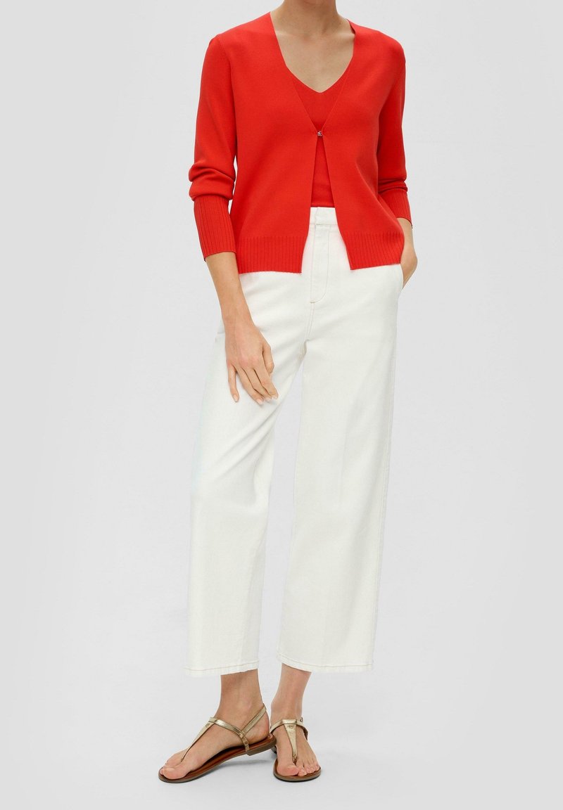 Femme porte un cardigan rouge vif à col en V sur un top assorti, pantalon blanc taille haute à jambes larges et sandales plates dorées, debout les mains dans les poches.