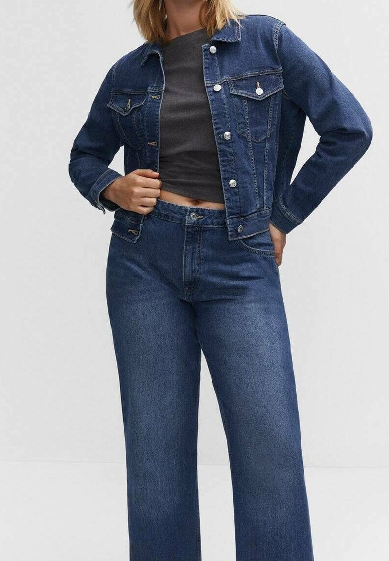 Femme portant une veste en jean bleu foncé et un jean large taille haute assorti avec un top court gris foncé.