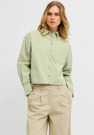 Frau mit schulterlangem blondem Haar, trägt ein hellgrünes kariertes Hemd mit Knöpfen und beige plissierte Hosen, Hände in den Taschen, neutraler Hintergrund.