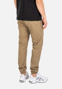 Mann trägt beige Jogginghosen und weiße Laufschuhe, steht mit dem Rücken zur Kamera vor einem einfarbigen Hintergrund.