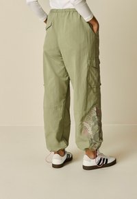 Pantalons cargo vert clair avec une taille élastique, des poches latérales et une broderie florale détaillée sur le bas de la jambe, associés à des baskets blanches.