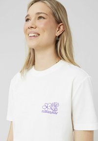T-shirt blanc avec un graphique floral violet et le texte « champ de fleurs » sur la poitrine gauche. Manches courtes, en coton, coupe décontractée.