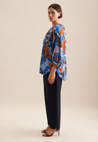 Blusa blu colorata con motivi floreali, caratterizzata da una vestibilità ampia, maniche a pallone e scollatura rotonda, abbinata a pantaloni neri e tacchi.