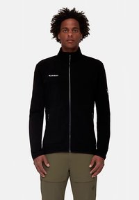 Fekete gyapjúdzseki magas gallérral, teljes hosszúságú elülső cipzárral és cipzáras oldalzsebekkel. A mellrészen fehér "MAMMUT" logó található.