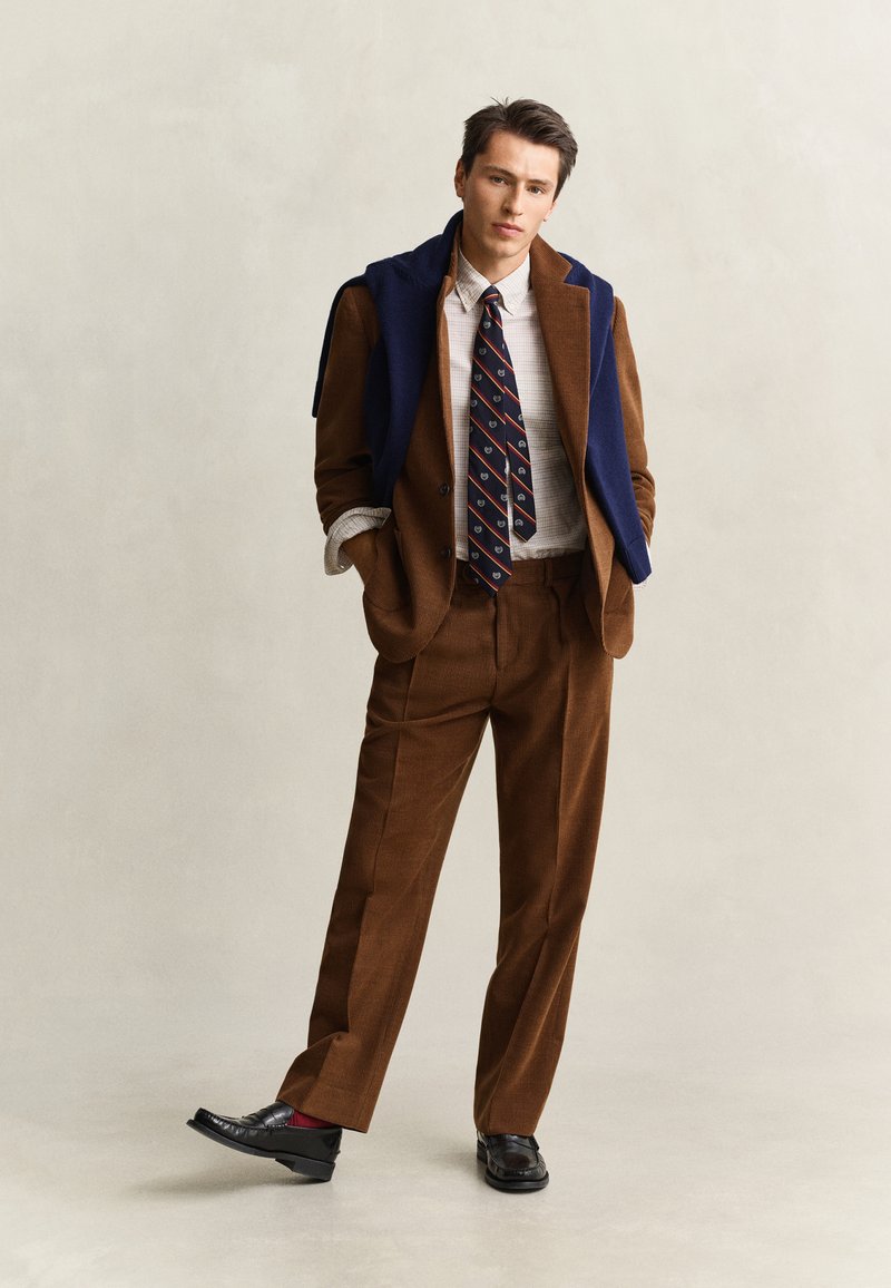 Homme portant un costume en velours côtelé marron avec un pull bleu drapé sur les épaules, une chemise blanche à carreaux, une cravate à motifs et des mocassins noirs.