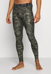Legginsy w wzór kamuflażowy w odcieniach zieleni, z szerokim czarnym pasem w talii z napisem "ARMOUR", wykonane z elastycznego, gładkiego materiału.
