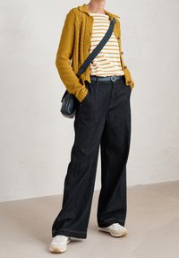 Person med senapsgul stickad kavaj, vit och senapsgul randig skjorta, mörka vida jeans, vita sneakers och axelväska.