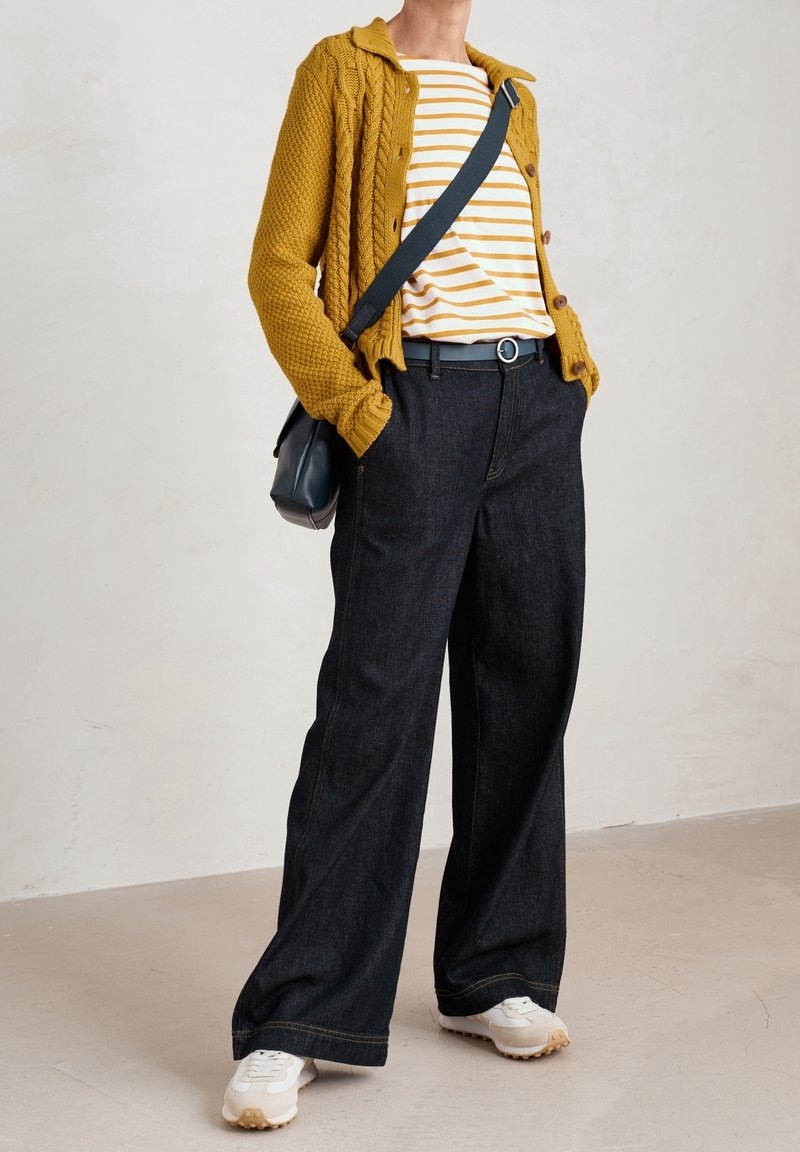 Person med senapsgul stickad kavaj, vit och senapsgul randig skjorta, mörka vida jeans, vita sneakers och axelväska.