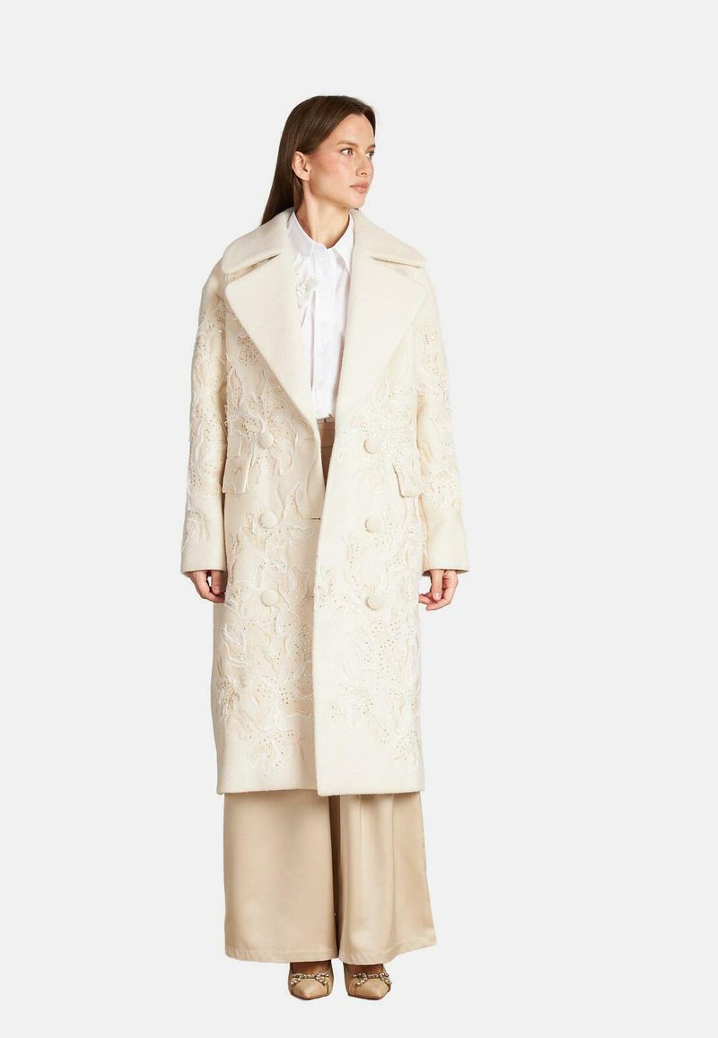 Largo abrigo crema con intrincados bordados florales, amplio cuello y cierres de botones, combinado con pantalones de pierna ancha beige y calzado adornado.