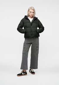 Melna pūkainā jaka ar kapuci, kas ir ar izšūtu dizainu un priekšējo rāvējslēdzēju. Pāris ar tumši pelēkiem kravas biksēm un melniem sporta apaviem.