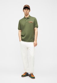 Άνδρας που φοράει ελιά πράσινο πόλο με λογότυπο αλόγου και ιππέα, άσπρο παντελόνι, πολύχρωμα παπούτσια και καφέ καπέλο, στέκεται με το ένα χέρι στην τσέπη.