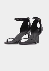 Paire de sandales noires à talons hauts avec bouts ouverts, fines brides à la cheville et petites boucles dorées sur un fond clair uni.