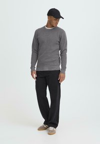 Szürke textúrájú pulóver kerek nyakkal, laza fekete nadrággal párosítva. A modell fekete sapkát és szürke sportcipőt visel, fehér csíkokkal.