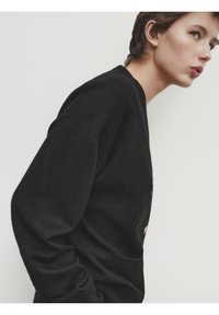Femme aux cheveux courts portant un pull noir à manches longues, penchée en avant sur un fond blanc uni.
