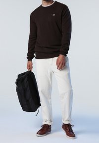 Pull en maille marron à col rond, porté avec un pantalon en velours côtelé blanc. Sac à dos noir dans une main, chaussures marron à lacets complètent la tenue.