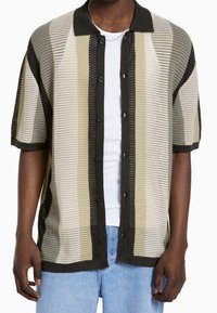 Camisa de manga corta con botones en un diseño de malla con rayas horizontales en beige, verde oliva y marrón oscuro. Presenta un collar oscuro y detalles acentuados.