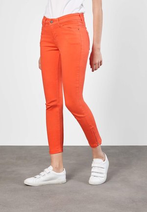 Persoon draagt slimfit feloranje broek met ritsen bij de enkels en witte sneakers met klittenbandsluitingen, staand op een grijze vloer.