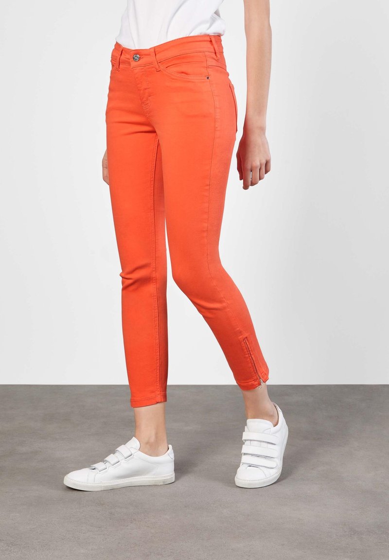 Personne portant un pantalon slim orange vif avec fermetures éclair aux chevilles et des baskets blanches à bandes Velcro, debout sur un sol gris.