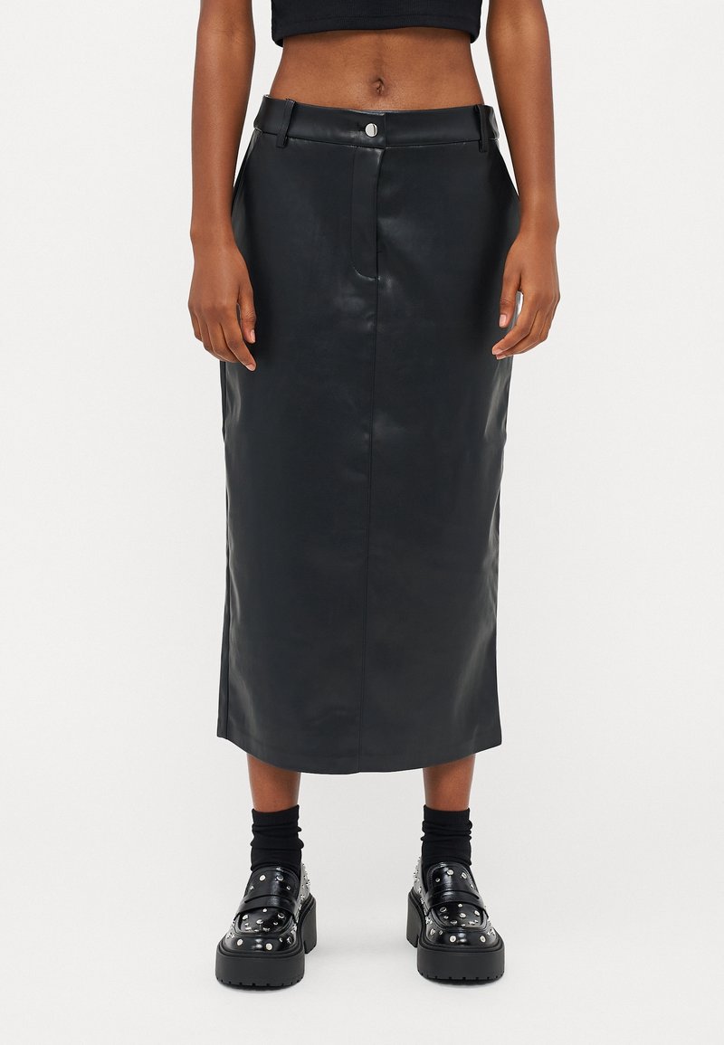 Jupe midi en cuir noir à la texture lisse, fermeture à boutons sur le devant et passants pour ceinture. Stylée avec des chaussures plateformes noires épaisses et des chaussettes.