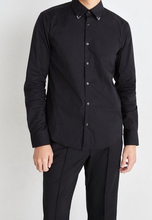 Homme portant une chemise noire ajustée à boutons avec manches longues et un pantalon noir, debout devant un fond blanc uni.