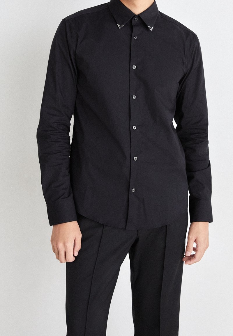 Homme portant une chemise noire ajustée à boutons avec manches longues et un pantalon noir, debout devant un fond blanc uni.