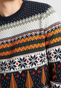 Stickad tröja med ett flerfärgat mönster på en mörkblå botten. Innehåller zigzag-, snöflinga- och trädmotiv i vitt, grönt, orange och rött.