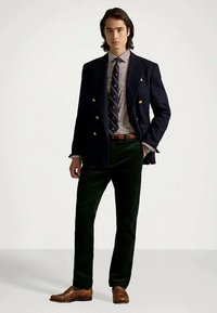 Blazer azul-marinho de abotoamento duplo, camisa xadrez, gravata às riscas, calças de veludo verdes e mocassins de couro castanho sobre um fundo simples.