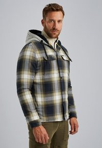 Chaqueta a cuadros negra, blanca y amarilla con capucha gris, con botones en la parte frontal y dos bolsillos en el pecho, fabricada con una suave mezcla de tejidos.