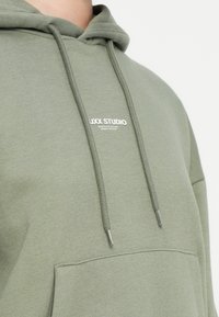 Katoenen hoodie in olijfgroen met een capuchon met trekkoord, een voorzak en een klein wit logo op de borst. Zachte textuur en een relaxte pasvorm.