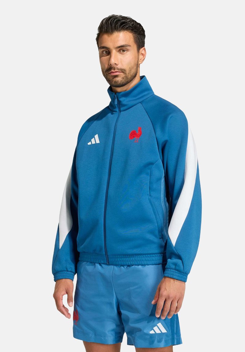 Modrá tréningová bunda s bielymi akcentmi, s logom červeného kohúta a pruhmi Adidas; v kombinácii s ladícími modrými šortkami. Mäkky, športový materiál.