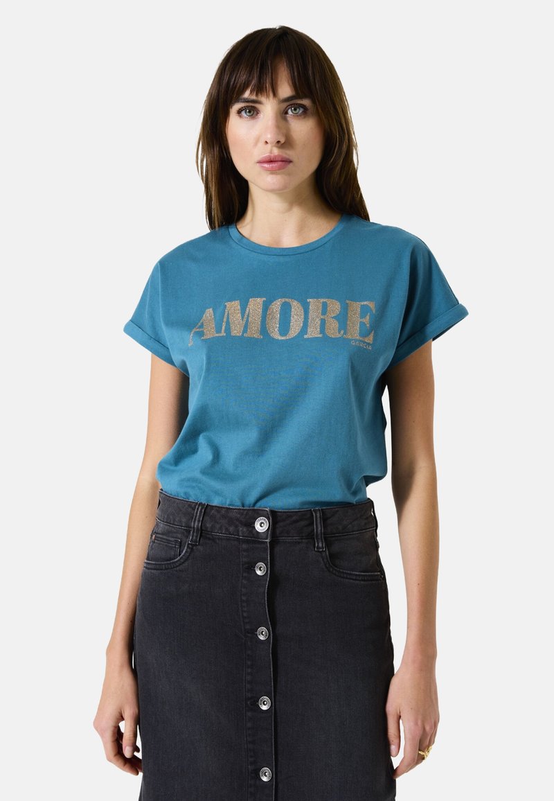 T-shirt turquoise avec des manches roulées arborant "AMORE" en paillettes dorées. Associé à une jupe en denim noire taille haute avec des boutons à l'avant.
